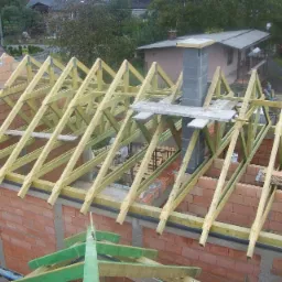 Wznoszenie konstrukcji dachu z drewnianych belek na murowanym budynku z cegły, z widocznym kominem w trakcie budowy i elementami deskowania.